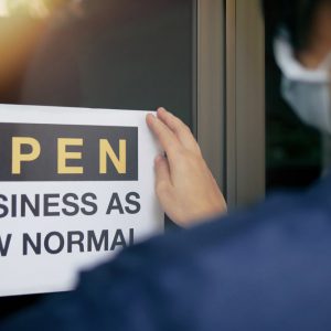 Rear view of business owner wearing medical mask placing open sign “OPEN BUSINESS AS NEW NORMAL” on front door.